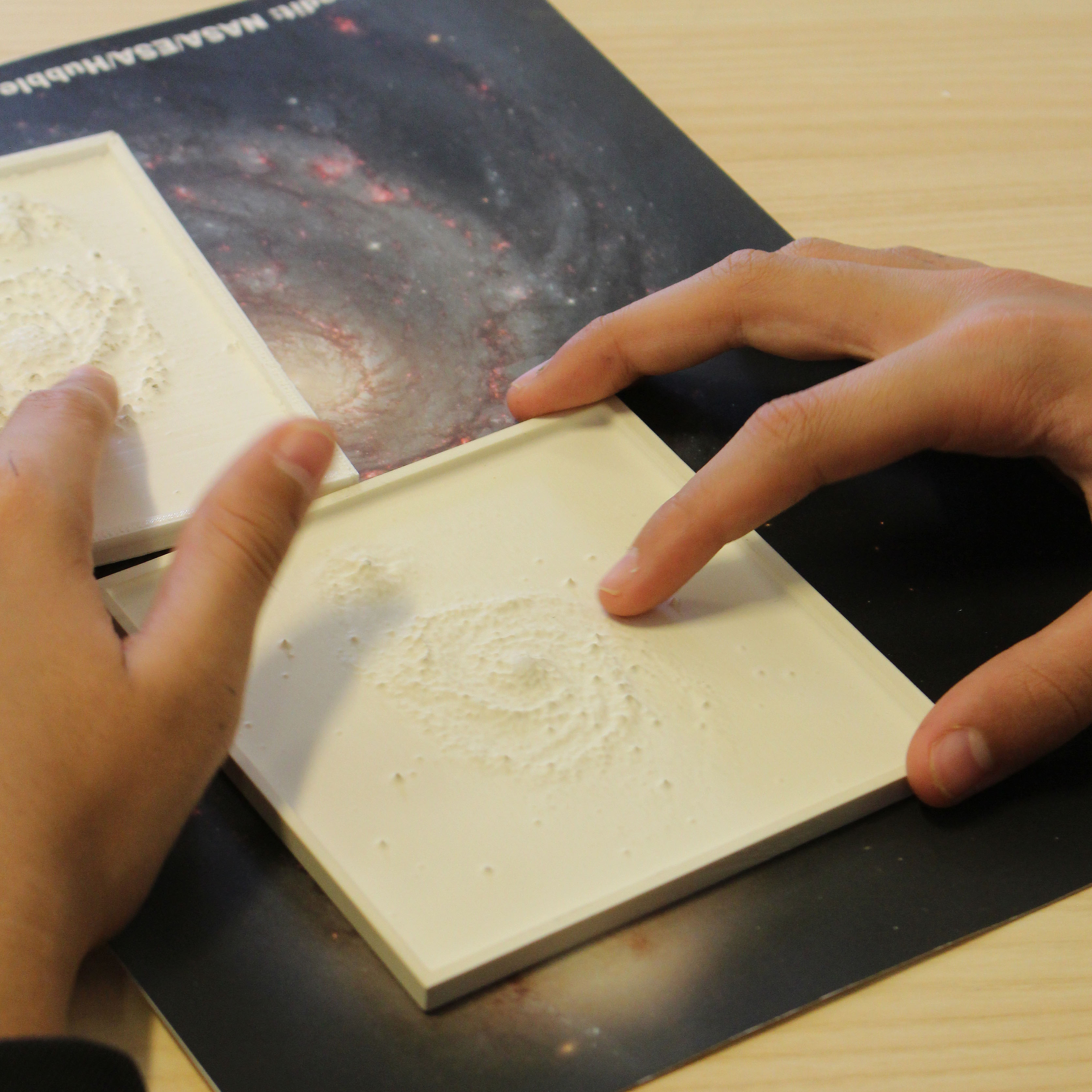 A student feels tactile images of galaxy Messier 51 with their fingertips. The tactile images are sitting on top of a full colour image of the same galaxy.